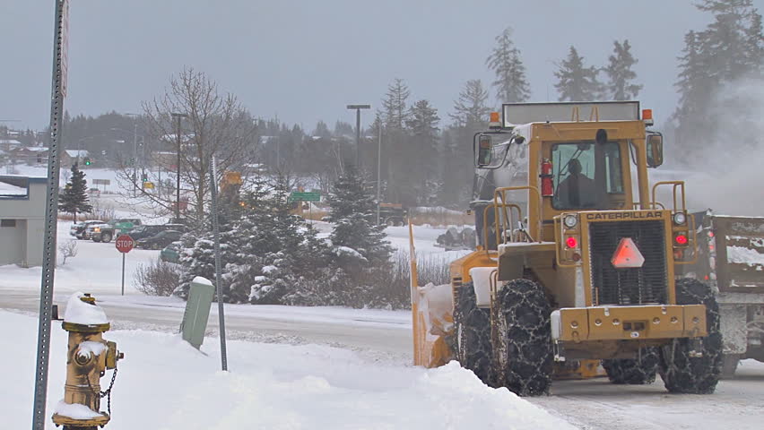HOMER, AK - CIRCA 2011: Snow blower on an excavator paired with a dump truck to clean plow piles on snowy city street.
