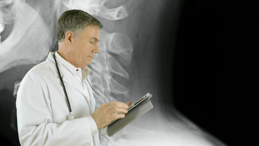 An x-ray image of a humane skull and cervical vertebrae are the backdrop for this physician who stops looking at the electronic tablet he is using and smiles for the camera.
