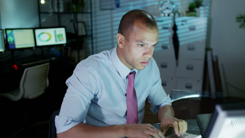 A young professional male is working late at night, a bank of computer screens can be seen in the background. He looks tired and uncomfortable and he loosens his tie