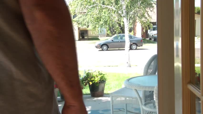 Man waits for car to pick him up by standing in front of glass door before light blue car pulls up, he leaves, and then enters car.