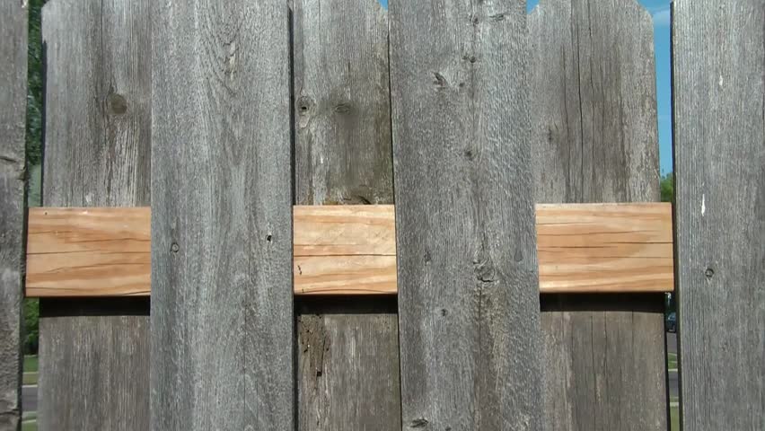 Clip of man in sunlight hammering in large nail into new wood board on fence.