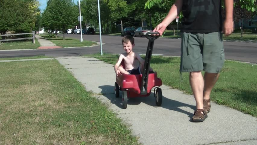 Little boy gets ride in red wagon with father pulling him down sidewalk occasionally smiling towards camera, in sun.