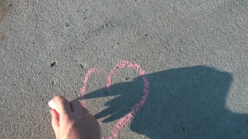 Using sidewalk chalk, a pink heart is drawn into driveway concrete on sunny day.