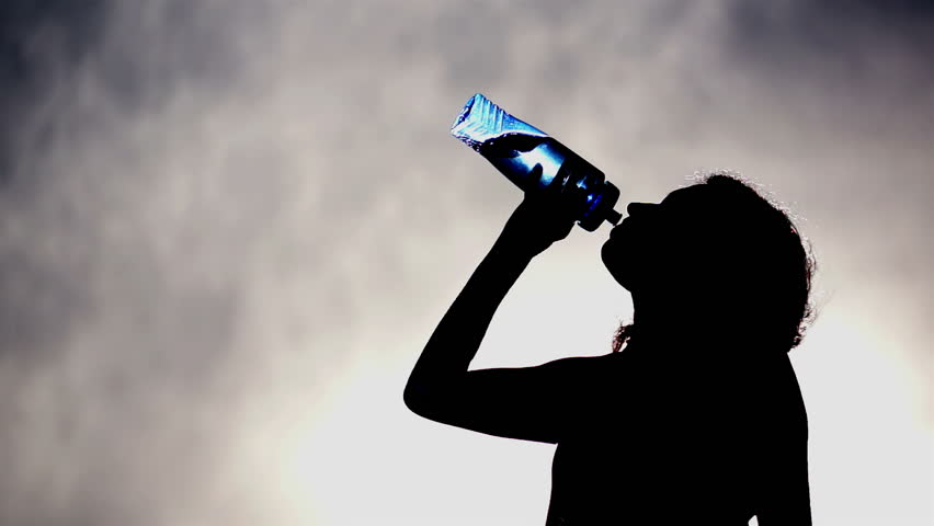 Silhouette of a woman drinking water out of a blue, translucent water bottle with a cloudy grey and blue sky background.