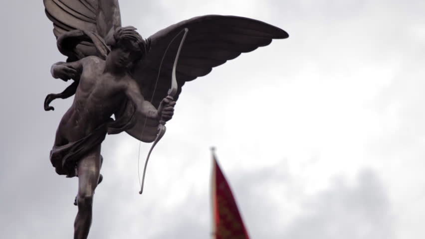 A stationary close-up shot of Eros statue on Piccadilly Circus in London. A flag is waving in the background, which is defocused. 