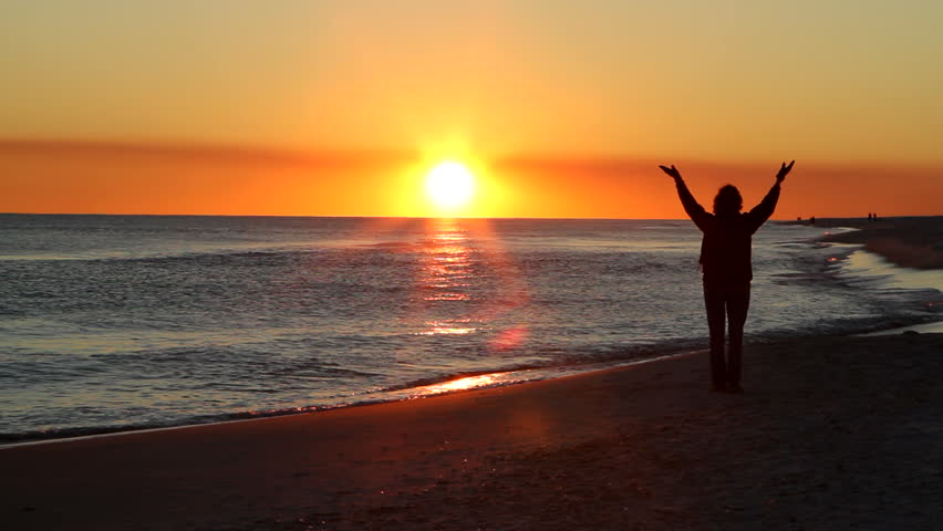 Woman raises her hands in the air to worship and pray at sunset at the seashore.