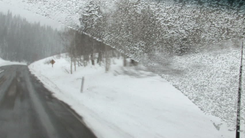 A blizzard in high mountains of central Utah. Driving car down a winding steep canyon looking through windshield. Snow and ice on road. Forest, trees and road. Camera moves with vehicle bounces.
