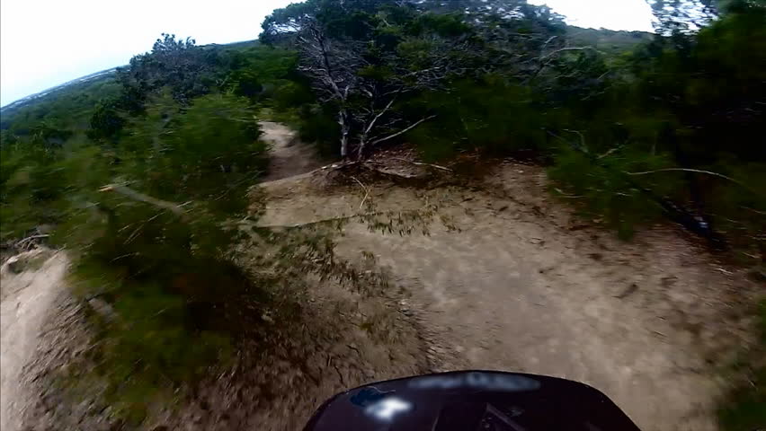 A POV, point of view, shot of a mountain biker riding down a path during the day. Another rider hits a jump in front of the camera and jumps over the trail.
