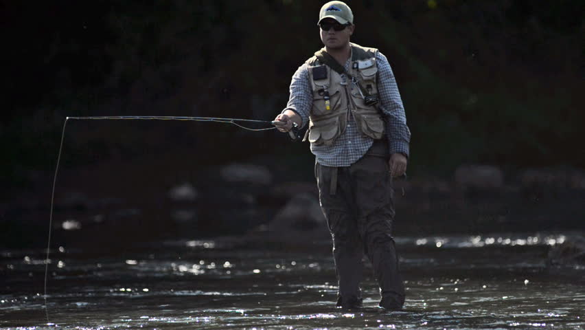 A man in waterproof overalls stands in a river and casts his lure with his rod while he spends his day fly fishing.