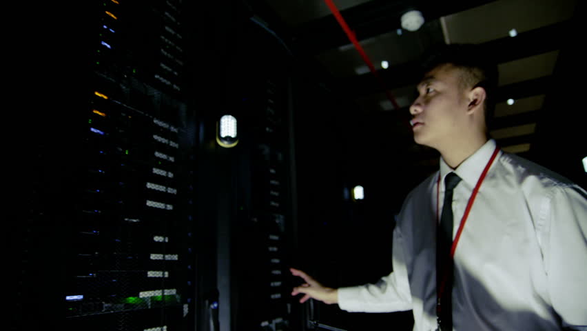 A young male IT engineer of Asian ethnicity is working in a data center with rows of server racks and super computers. He is looking into cabinets and checking cables and other equipment. Slow motion.
