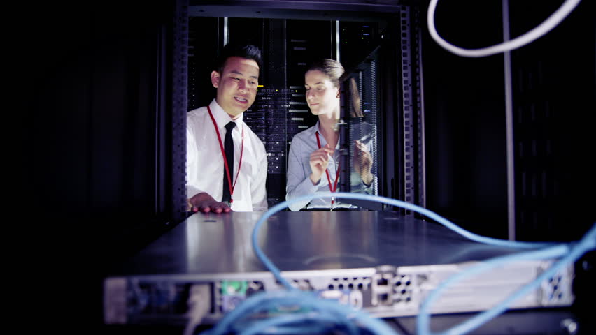 Male and female IT engineers are working in a data center with rows of server racks and computers. They are discussing their work as they check cables and other equipment. In slow motion.
