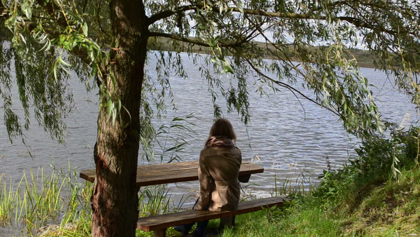 woman girl sit on wood bench under willow tree branch move in wind near lake. recreational recess solitude place.