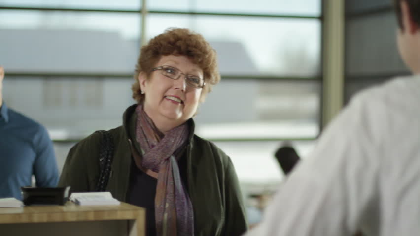 Female customer finishes her transaction, leaves and male customer comes up to bank teller. Over the shoulder shot from behind teller showing other customers and bank interior. 