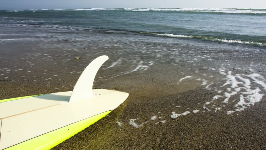 A time lapse of waves slowly washing up on a surfboard laying the sand.