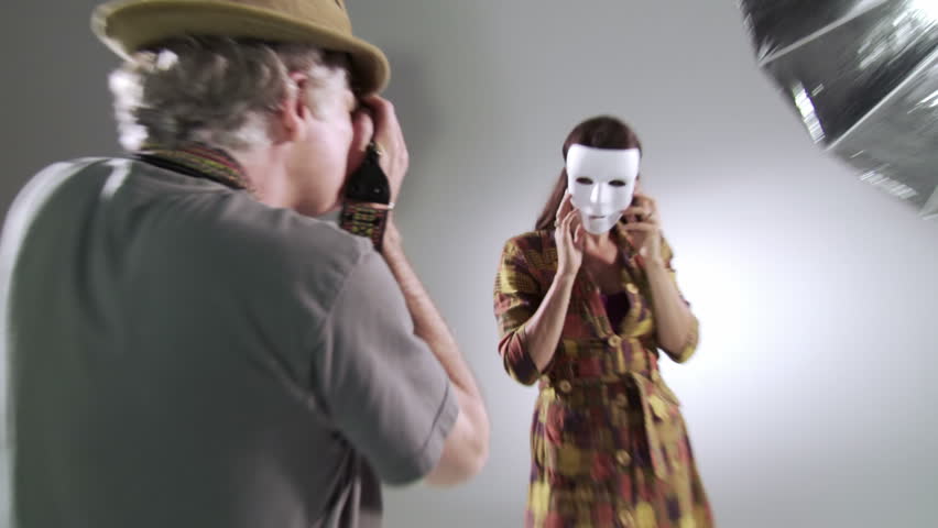Photographer tries to reveal the person behind the mask as he takes pictures of female model Edgy, hand-held camera in studio.