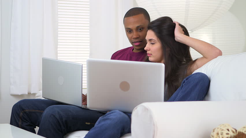 African American and Caucasian couple using laptop computers on couch