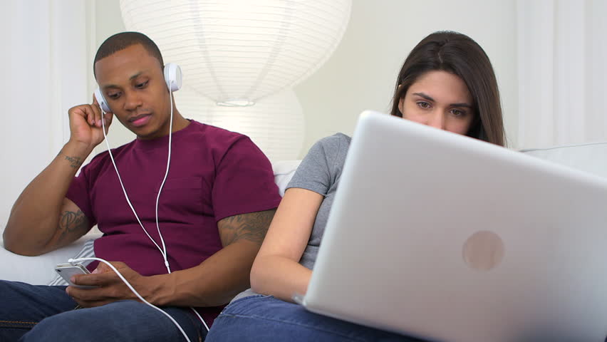Black man listening to music on smart phone then stopping to kiss girlfriend using laptop computer