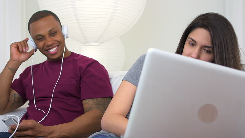 Black man listening to music on smart phone while girlfriend uses laptop computer