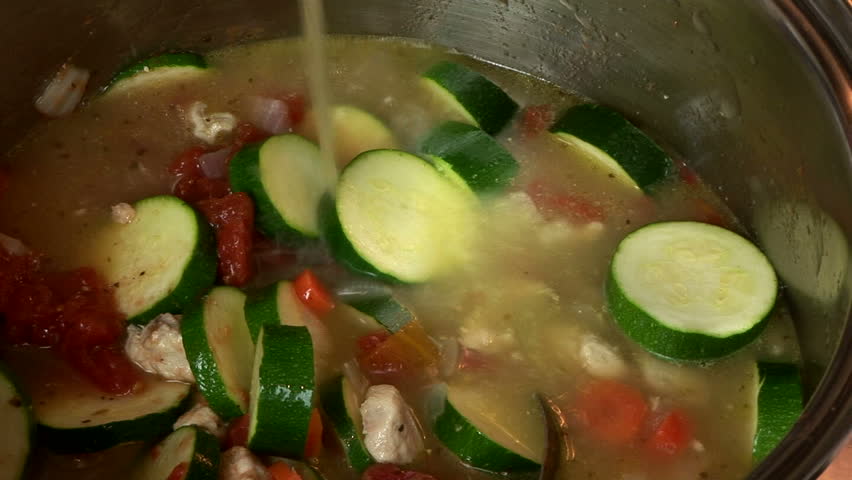 A cook adding chicken broth to a large pot of soup vegetables