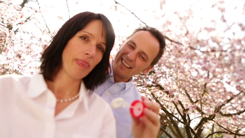 Attractive mature woman blowing soap bubbles in slow motion with her husband, the happy couple laughing under cherry blossoms 