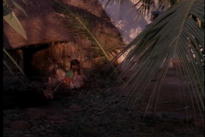 INDIA - DECEMBER 16, 1988: Camera pans from woman chopping sticks outside hut, up to top of solar powered street light.