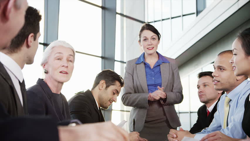 A confident and happy business team of mixed ages and ethnicity are holding a meeting in a light, modern office building. They applaud a comment from one of the group 