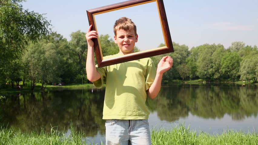 Young boy holds picture frame and imitates leaf over frames near pond in park at summer sunny day