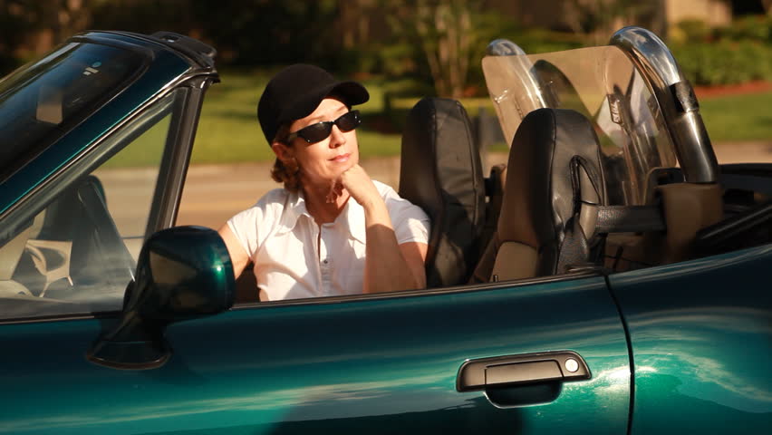 A woman sitting in a convertible sports car smiles at a man who is getting into the vehicle with her.