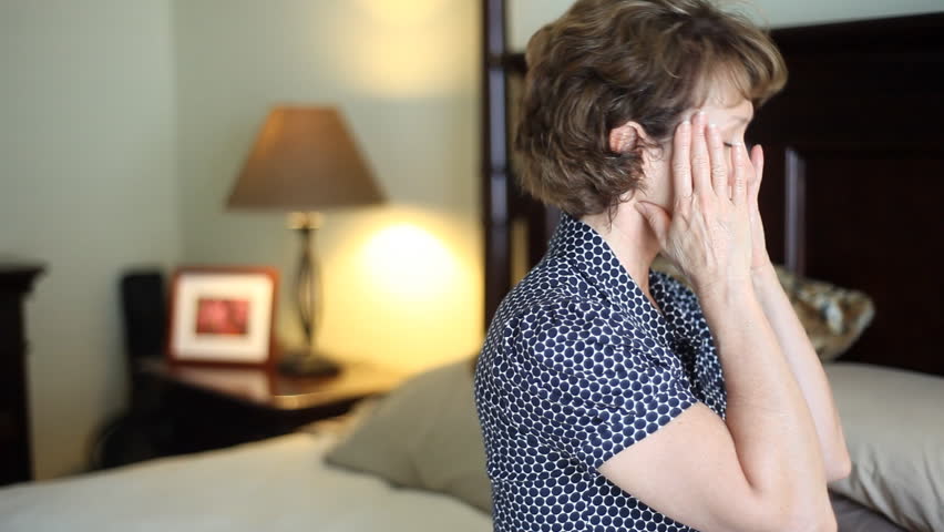 A troubled, stressed out woman sitting on her bed who appears to be suffering from a headache