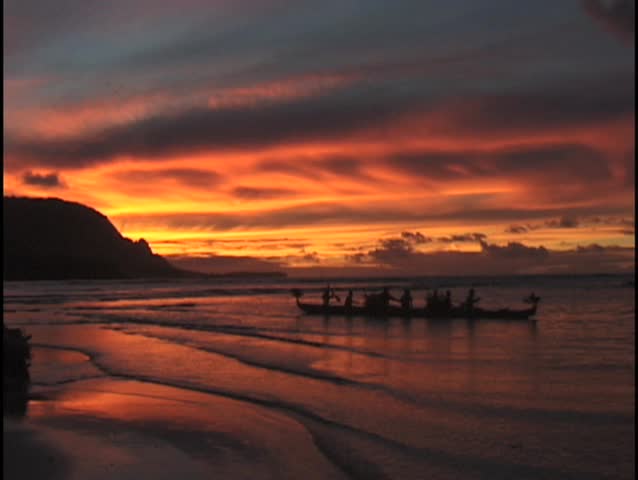 Fire red sunset silhouette of Hawaiian outrigger boat with crew and distant  mountain range.