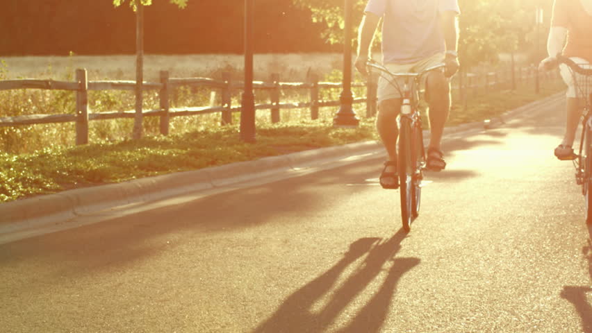 An older couple rides bikes down the street at sunset in slow motion as the camera follows them. With lens flare