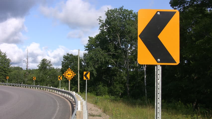 Sign at road corner. One of several arrow signs indicating a sharp corner in the road. Van drives by towards end of clip. Ontario, Canada. 