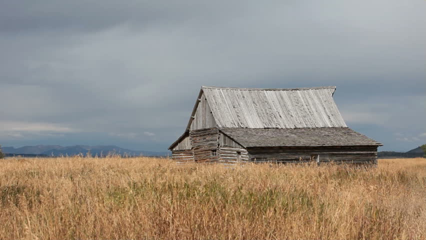 An old wood log barn in the valley meadow of Grand Teton National Park Wyoming. Grass blowing in the wind. Slow pan. Clouds cover top peaks of the mountain range. Pioneer building on Mormon Row.