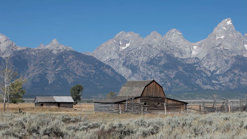 Old ranch wood log barn and sheds in valley meadow of Grand Teton National Park Wyoming. Grass blowing. Slow pan. Clear blue sky above peaks of the mountain range. Pioneer building on Mormon Row.
