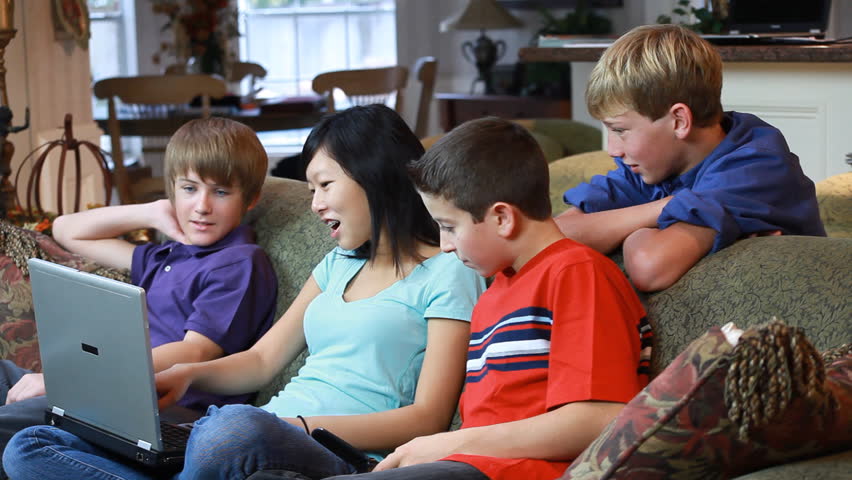 A pretty Asian teenager sitting on a couch, and holding a laptop engages in conversation with a group of her friends.