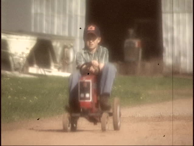 Slow motion - Vintage film look of a Rural farm boy (approximately 8 yrs. old) with a tractor pedal toy heading to the barn.