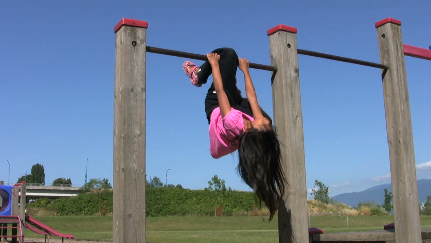 A cute little Asian girl shows how she can hang upside down from the monkey bars at the playground during summer.
