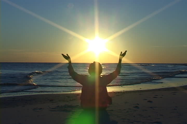 Mature woman sits on the beach with her hands raised in prayer at sunset. Shot on miniDV from a tripod.