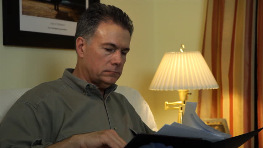 A man sitting in his living room looking through some paperwork stops and smiles at the camera.