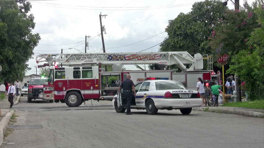 Video of fire trucks and police car at a residence home fire. Neighbors and new reporters walking around. Firemen relaxing after fire is put out. 