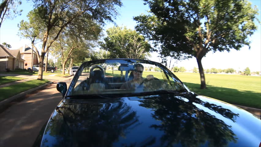 A man in a convertible sports car, drives down a tree lined street and pulls into his garage. Fisheye