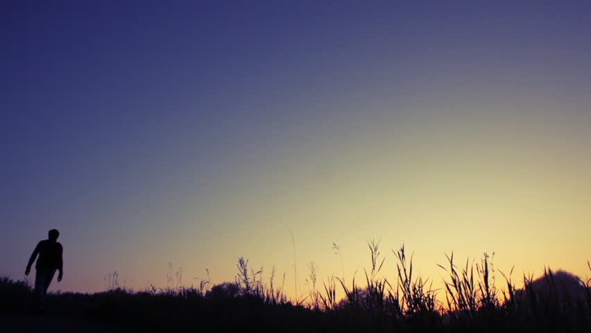 silhouette of man with rucksack walks uphill against sunset sky from camera 