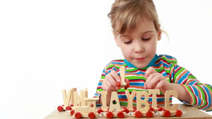 Little girl in a multicolored striped shirt with long sleeves concatenates trailers of train, consisting of letters of alphabet, front 