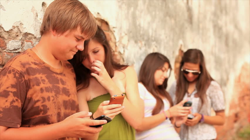 A group of teenagers hanging out by an old brick wall, using cell phones to send text messages and talking to each other.