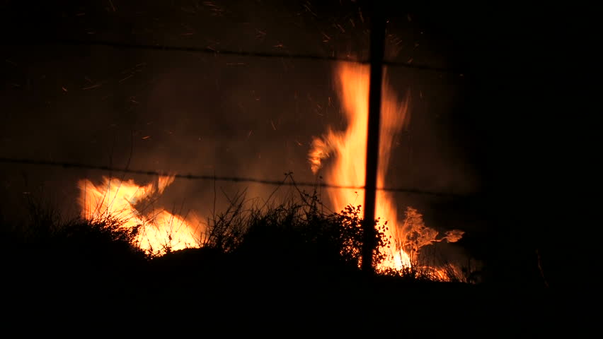 During a California wildfire, brush is consumed by fire. The blaze grows as firemen rush to save homes. You can hear a bulldozer nearby clearing the hillside.