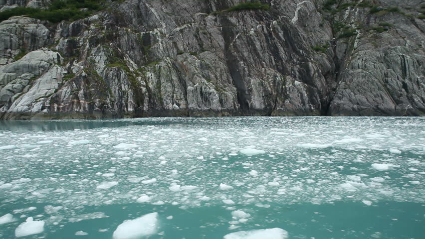 Ice flow and ice bergs floating and waterfalls down sheer cliffs as ship passes in glacial bay in Kenai Peninsula, Alaska. Steep rock ledges fall into ocean fjord leaving ice chunks.