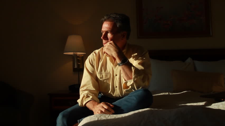 A handsome, mature man bathed in warm afternoon sunlight coming through his hotel window contemplates something troublesome.