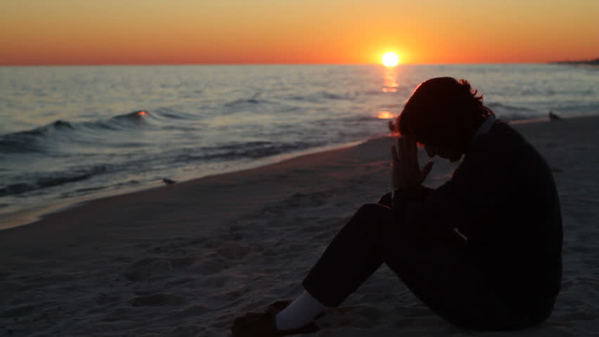 Mature woman sits on the beach praying as the sun sets over the Gulf of Mexico in the background.