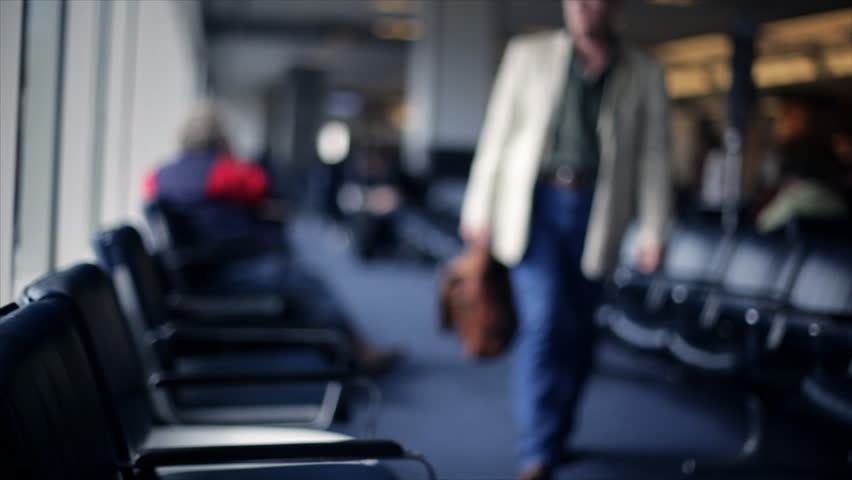 A man in an airport carrying a briefcase finds a seat by a large window then checks the time