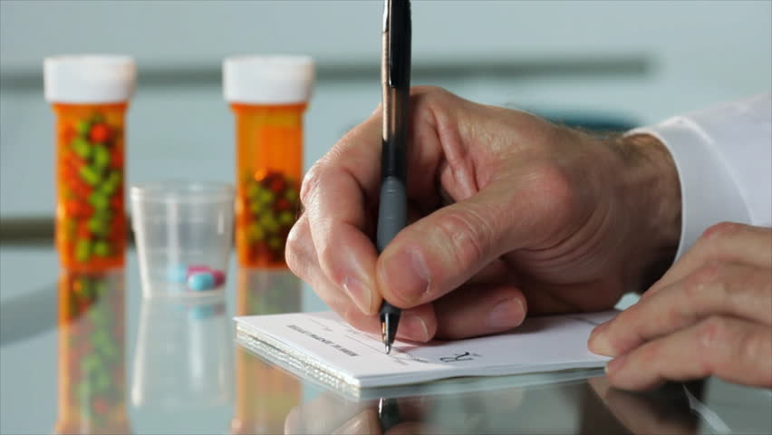 A close up of a male physician's hand writing out a prescription with out of focus pharmaceuticals pill bottles in the background.